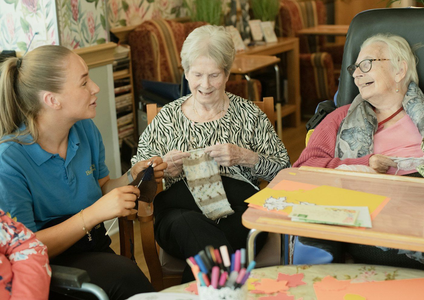 A caregiver helping two elderly residents with a knitting activity